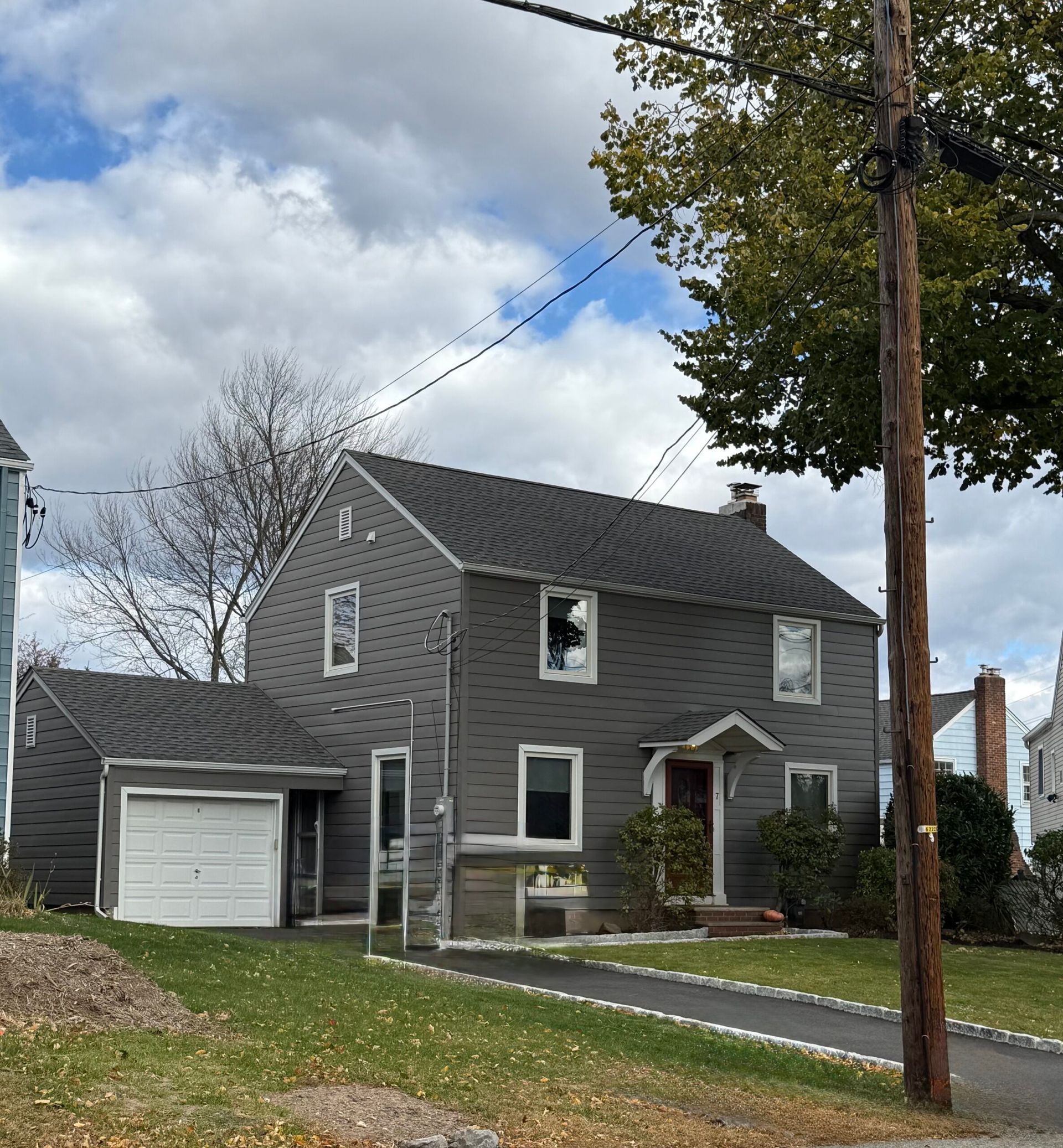 Gray two-story house with attached garage, driveway, and lawn. Cloudy sky, utility pole in foreground.