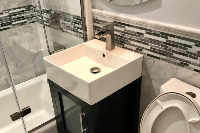 Small bathroom with a square sink on a dark cabinet, toilet, tiled wall, and a bathtub with glass enclosure.