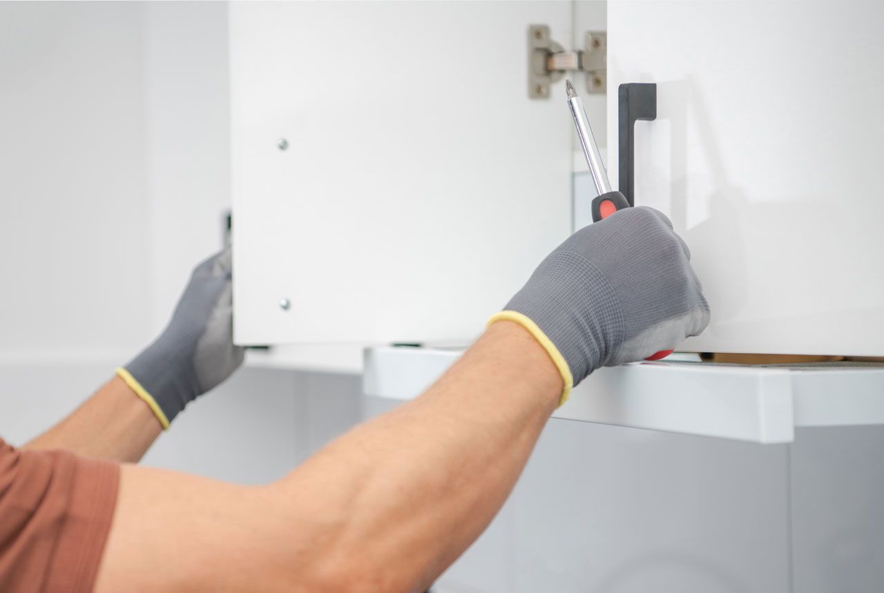 Person wearing gloves uses screwdriver to adjust cabinet hinge.