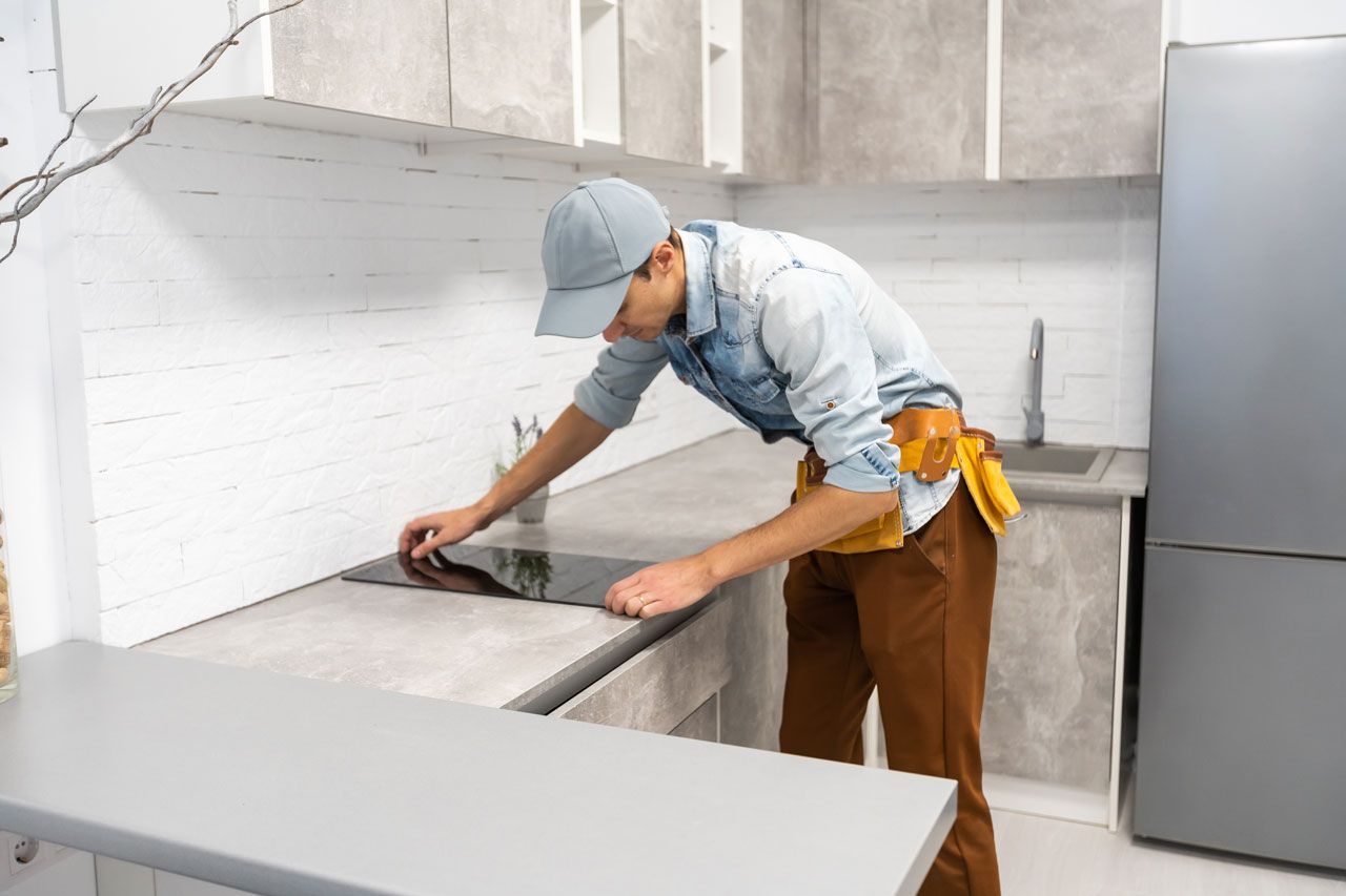 Person installing cooktop in a modern kitchen, wearing cap, tool belt, and brown pants.