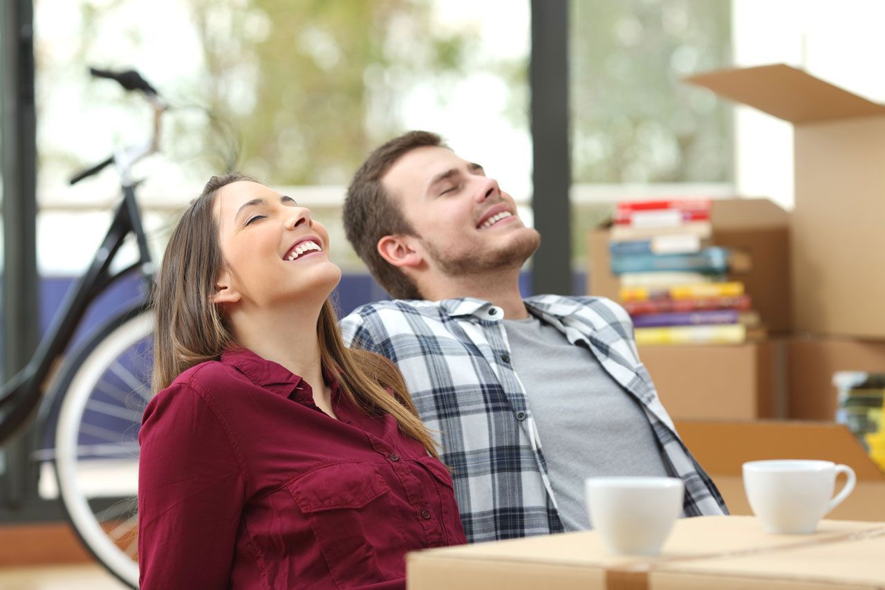 Couple smiles, relaxing in new home with boxes, bike, and mugs.