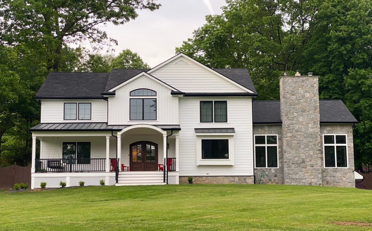 White house with black roof, front porch, and stone chimney set on a green lawn.