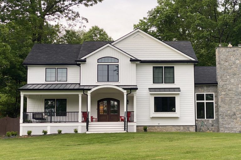 White farmhouse with dark roof, black-framed windows, stone chimney, and front porch, set on green lawn.