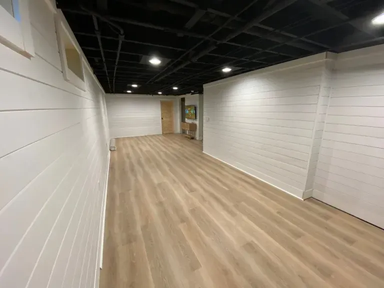 A long, rectangular finished basement with white paneled walls, a black ceiling, and light wood-look flooring.
