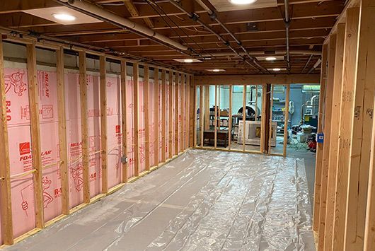 Basement under construction, with wooden framing, pink insulation, and a plastic floor covering.