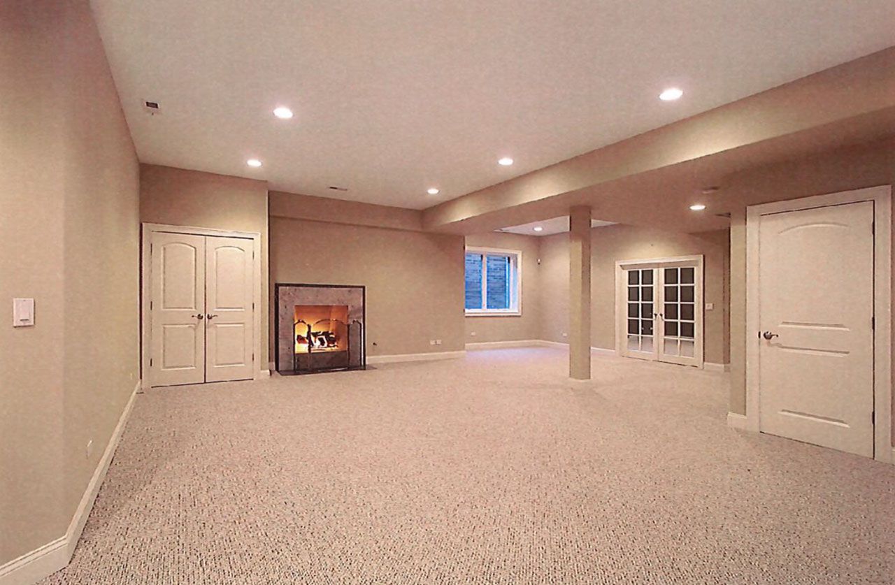 Empty basement room with fireplace, doors, window, and beige carpeting.