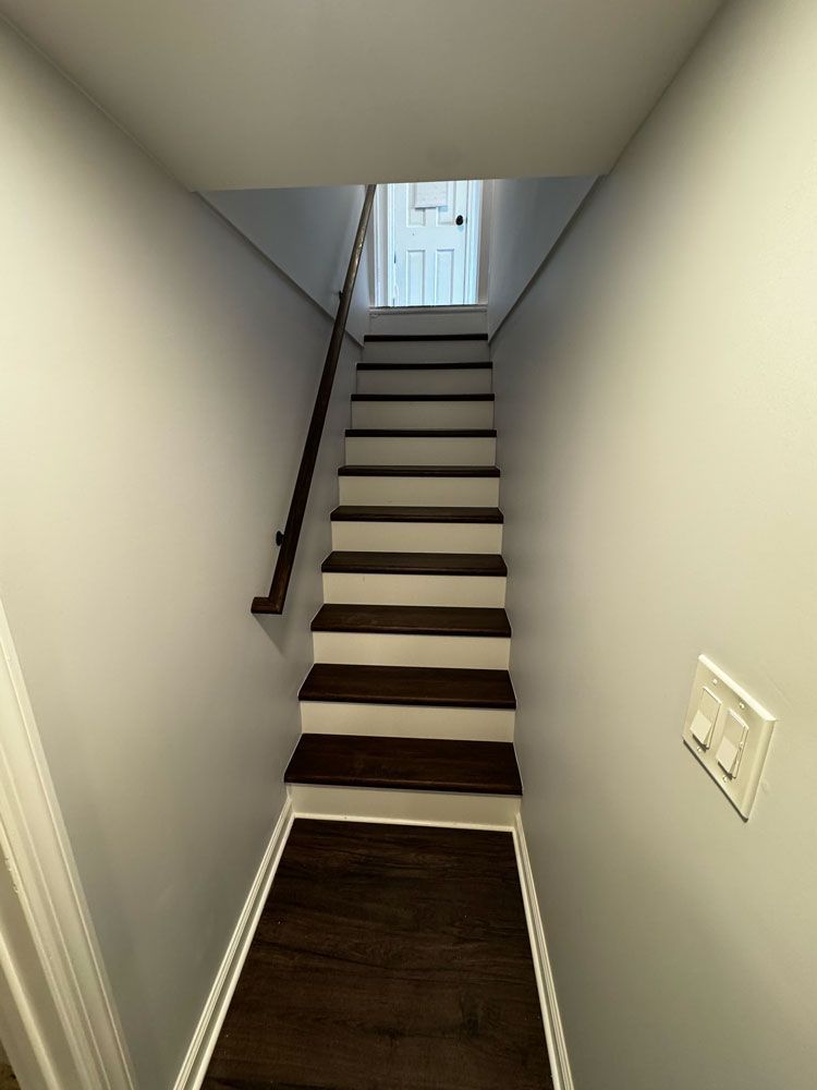 Narrow staircase leading up with dark wood steps and white walls. Handrail on the left.