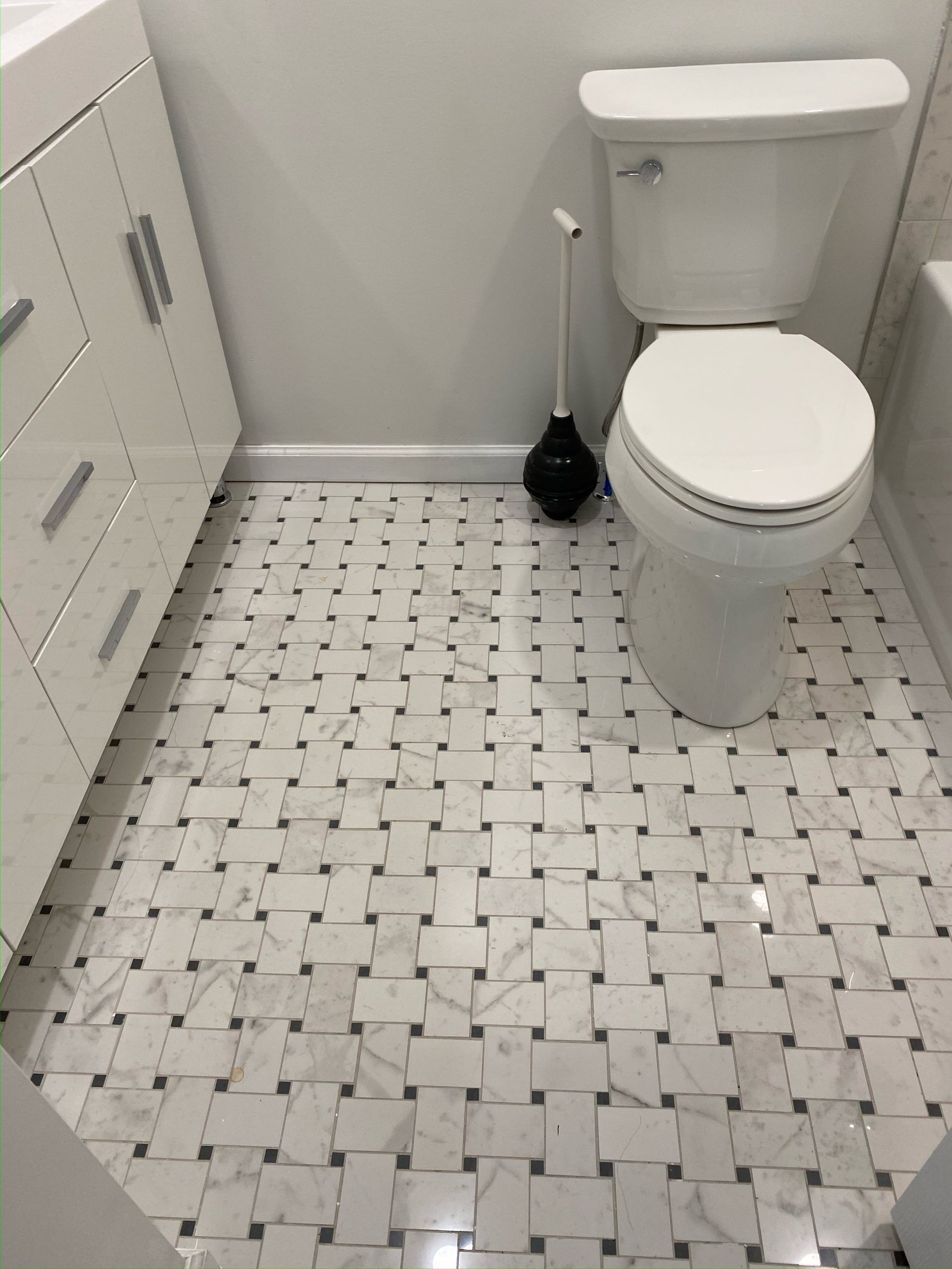 Bathroom with white vanity, toilet, and patterned tile floor. A plunger stands next to the toilet.