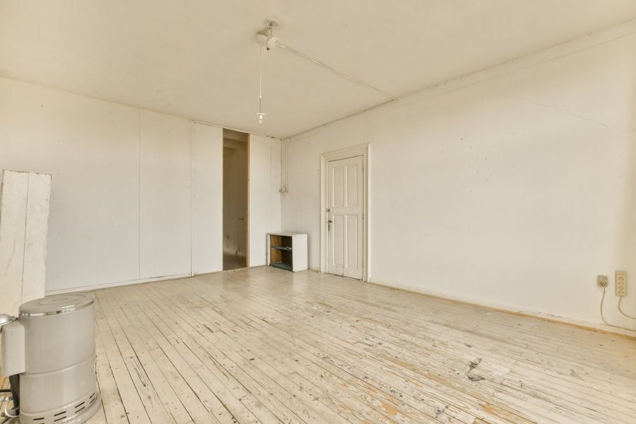 Empty, white-walled room with wood floor. Contains a door, open doorway, and a metal drum.