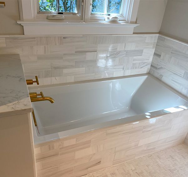 White tiled bathroom with a bathtub, gold fixtures, and a window.