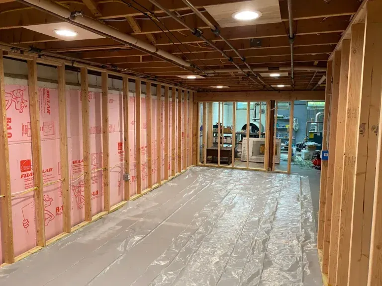 Framed basement room, insulation visible, plastic flooring, doorway to another space with light.