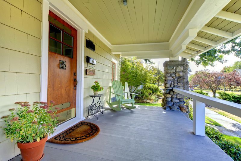 Beige house porch with a wooden door, potted plants, and green rocking chairs.