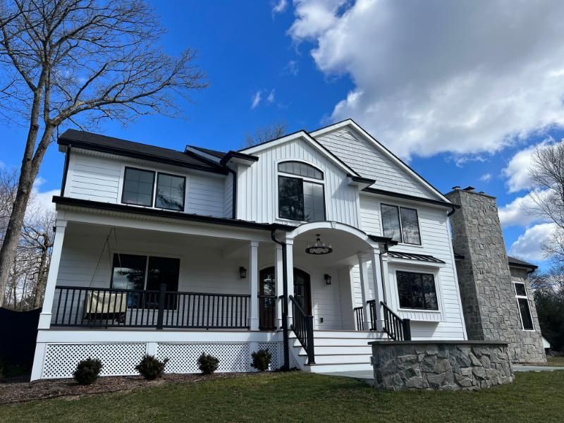 White two-story house with black trim, a covered porch, and a stone chimney under a blue sky.