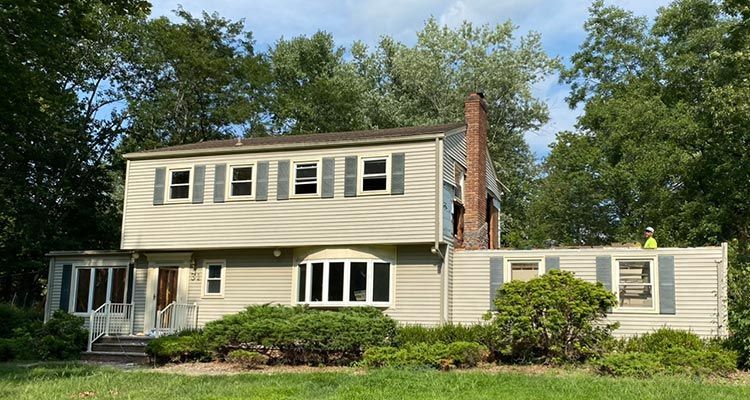 Two-story beige house with gray shutters and a red brick chimney, surrounded by green trees and bushes.