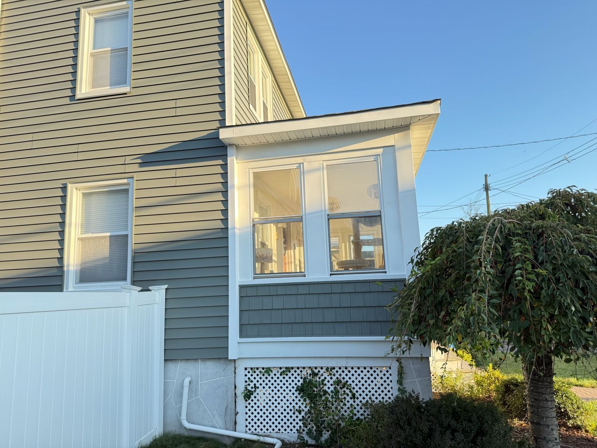 Gray-sided house with a small windowed porch, a white fence, and a small tree against a blue sky.