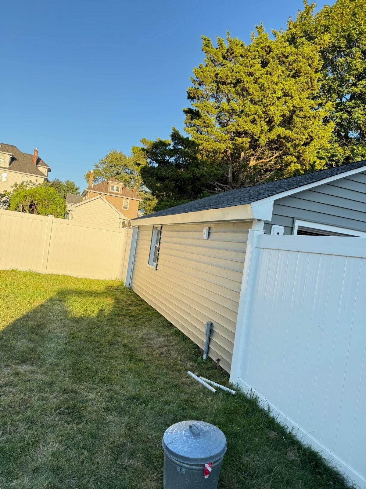 Tan-sided building with white fence, green lawn, and blue sky. A metal trash can sits in the foreground.
