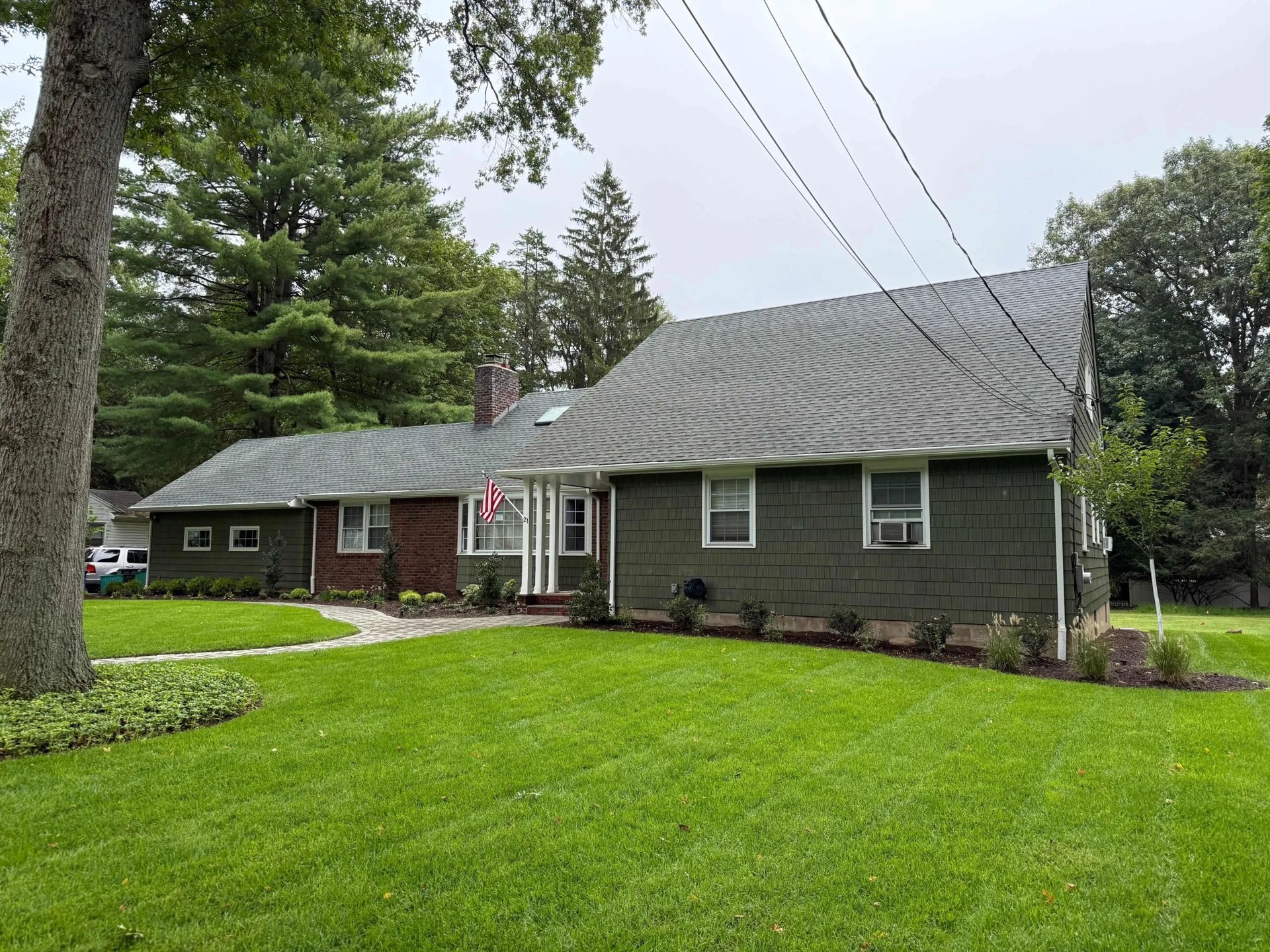 Green house with gray roof, brick and green siding, set on green lawn with trees in the background.