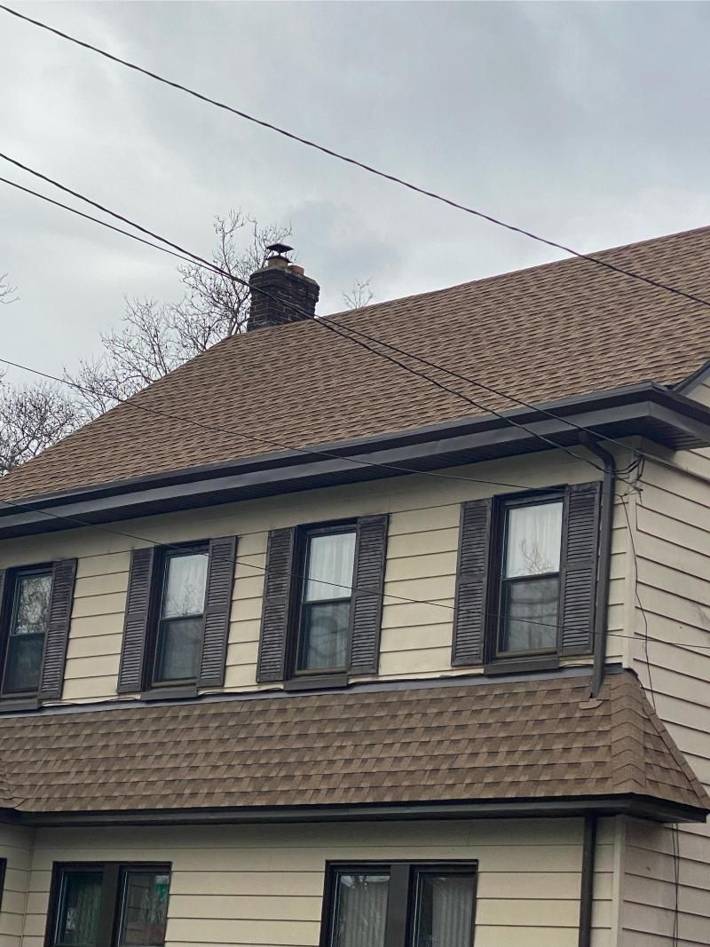 Two-story house with brown roof and shutters, chimney on roof, cloudy sky.