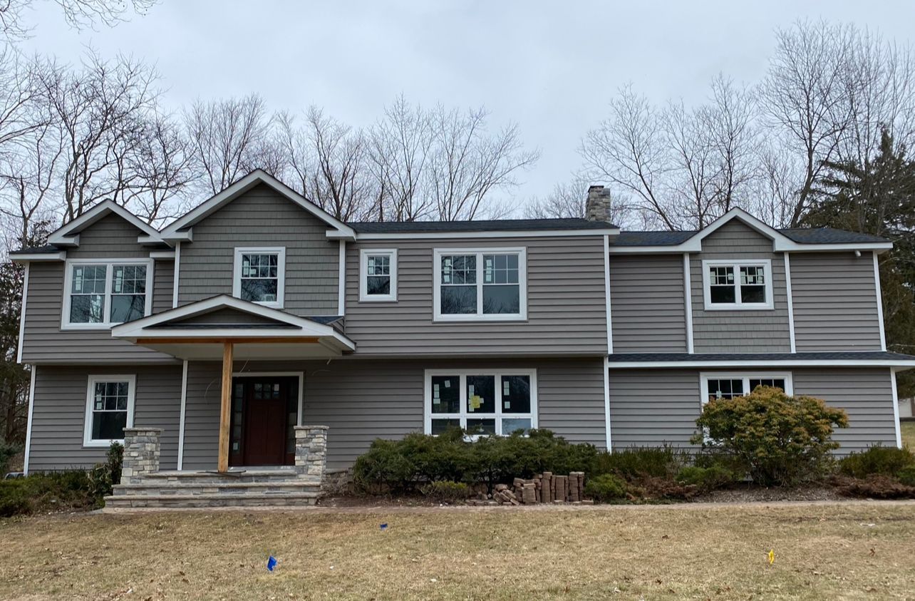 Two-story gray house with brown front door, stone steps, and windows; bare trees in the background.
