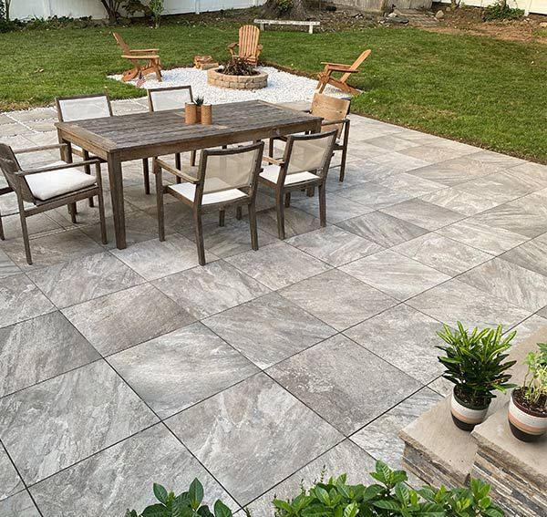 Outdoor patio with gray stone tiles, dining table, chairs, and fire pit on grass.