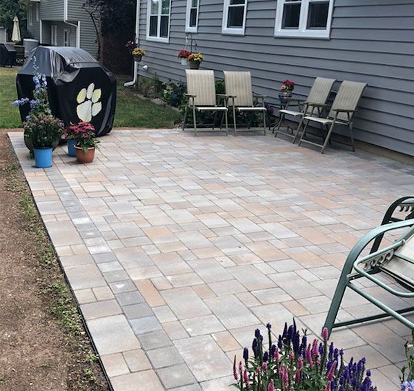Paver patio with chairs, a grill, and flowers next to a gray house.