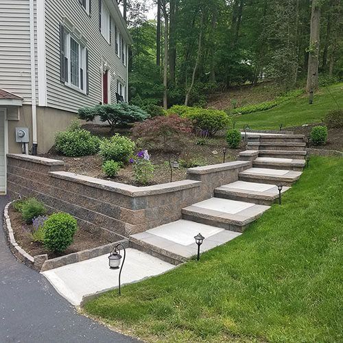 Stone staircase and retaining wall leading up to a house with landscaped garden beds, lush grass and trees.