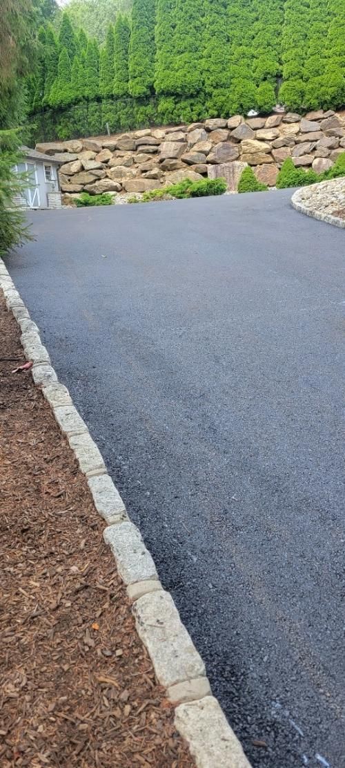 Asphalt driveway edged by stone and mulch, leading to a stone wall with greenery and trees.