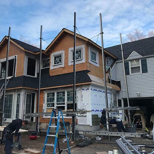 House under construction with scaffolding. Workers, plywood, blue wrap, ladder, and black roofing.