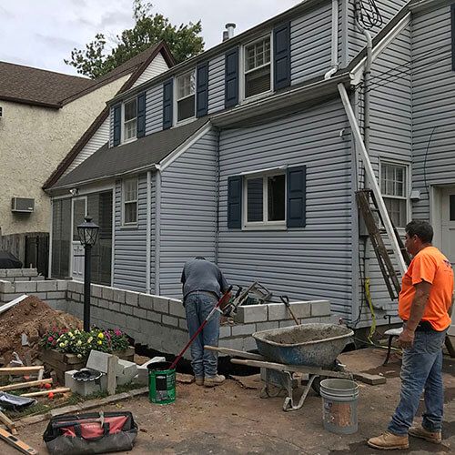 Construction workers building a brick wall outside a two-story blue-sided house.