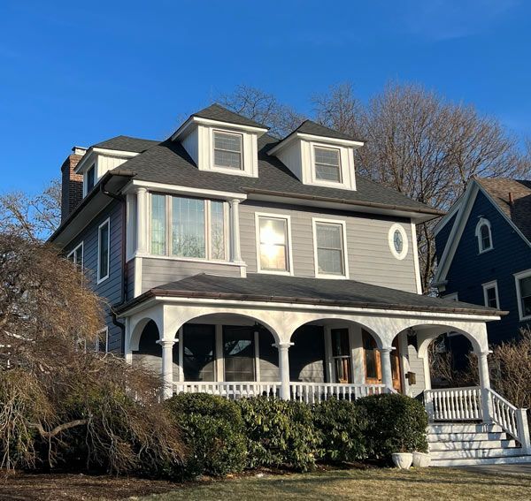 Two-story gray house with porch and dormers, blue sky, tree in the front yard.