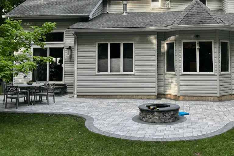 Backyard patio with fire pit, table, and house with windows and gray siding.