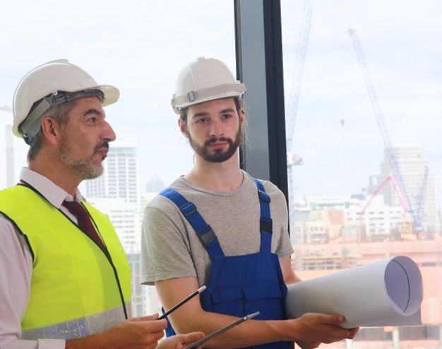 Two construction workers in hard hats discussing plans by a window overlooking a building site.