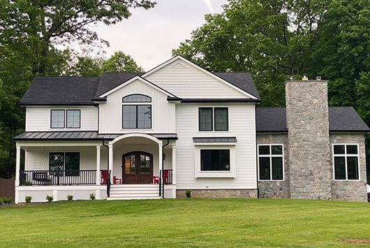 White two-story house with black roof, front porch, and stone chimney. Green lawn and trees in background.