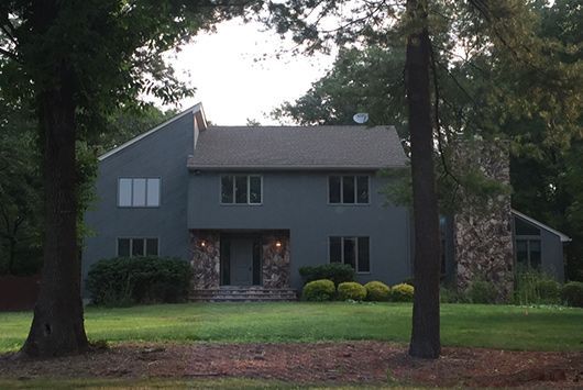 Two-story gray house with stone chimney and entrance, surrounded by trees and a grassy yard.