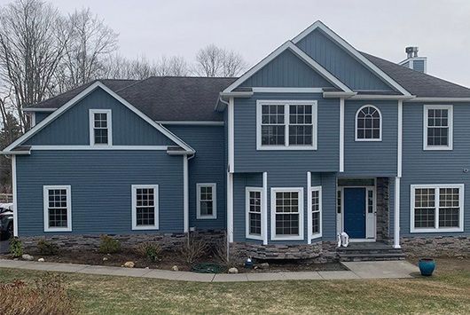 Blue two-story house with white trim and dark gray roof. Stone foundation, blue front door, and gray sky.