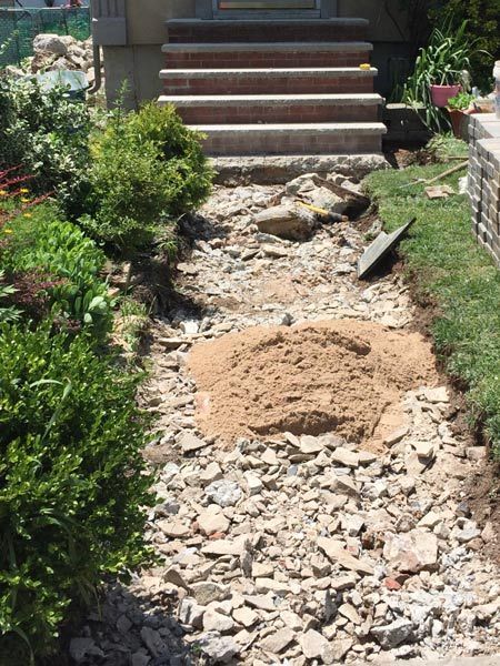 A walkway to a front door is under construction, with a pile of sand and broken debris. Steps lead up to the door.