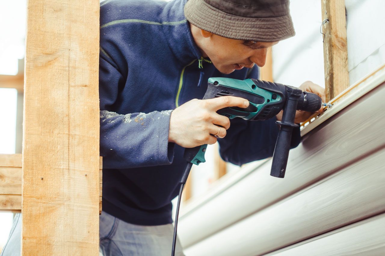 Man drills into siding on a building's exterior, wearing a blue jacket and a hat.