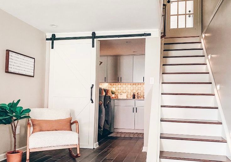 White barn door opens to a kitchen, beside a staircase with white steps. A chair and plant are in the foreground.