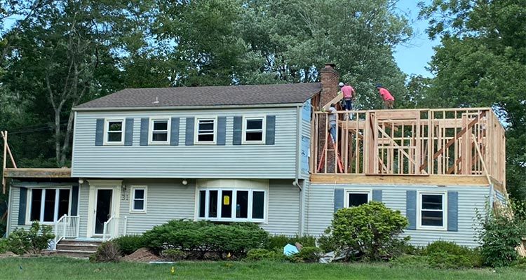 House under construction; workers on roof and framing a second-story addition; gray siding, green trees.