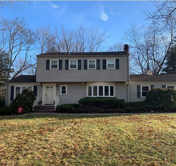 Two-story house with light gray siding, dark shutters, and brown roof on a grassy lot.