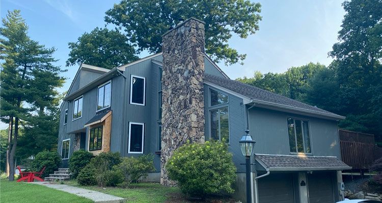 Gray house with stone chimney, windows, and surrounding trees.