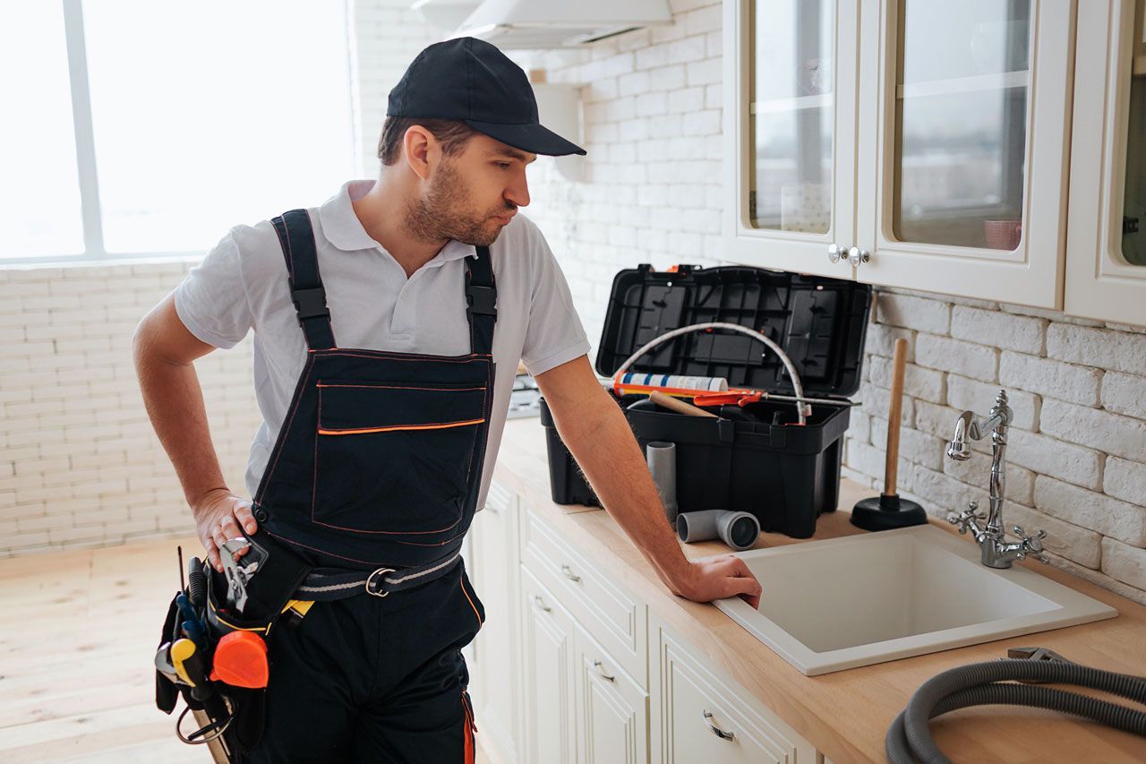 Plumber in work clothes leaning on kitchen counter, tools beside sink.
