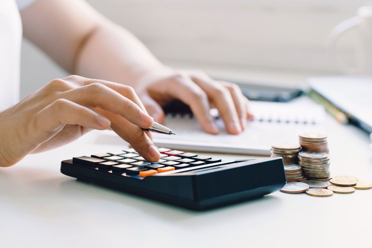 Person using a calculator with stacks of coins and a notepad on a white table.