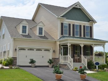Two-story beige house with a porch and garage, green shingles, and a paved driveway.