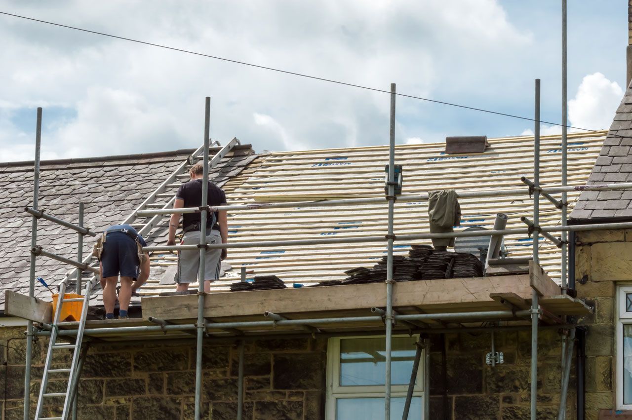 Two roofers on scaffolding, working on a tiled roof. Cloudy sky in the background.