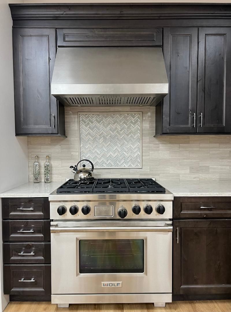 Stainless steel oven and range hood in a dark wood kitchen with light tile backsplash.