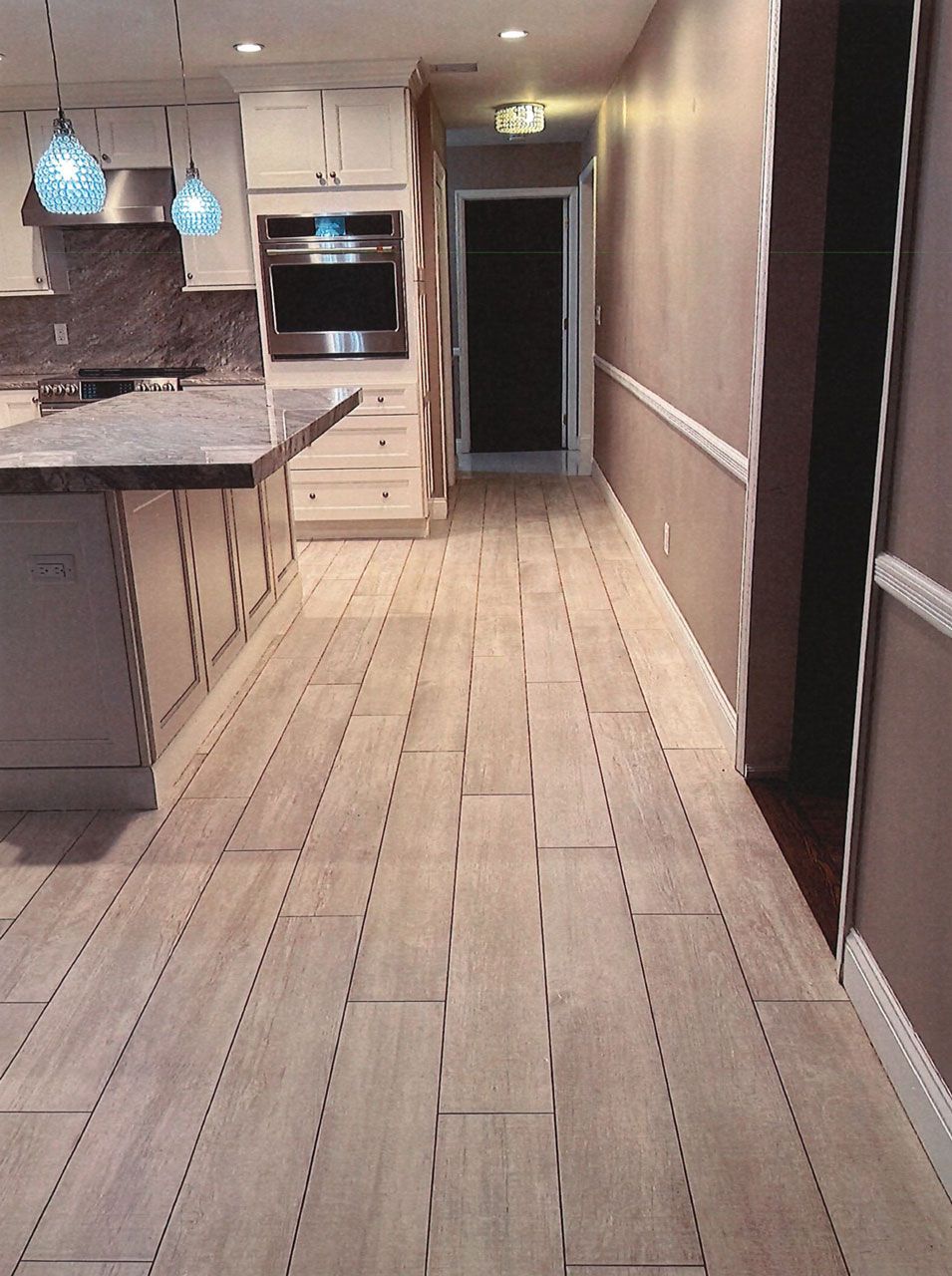 Kitchen with light wood-look flooring and a hallway leading to a dark door; built-in oven, countertops, and overhead lights.
