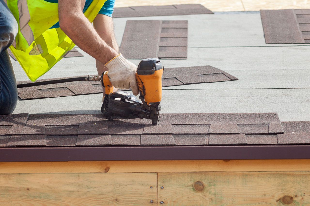 Roofer using a nail gun to install brown asphalt shingles on a roof.