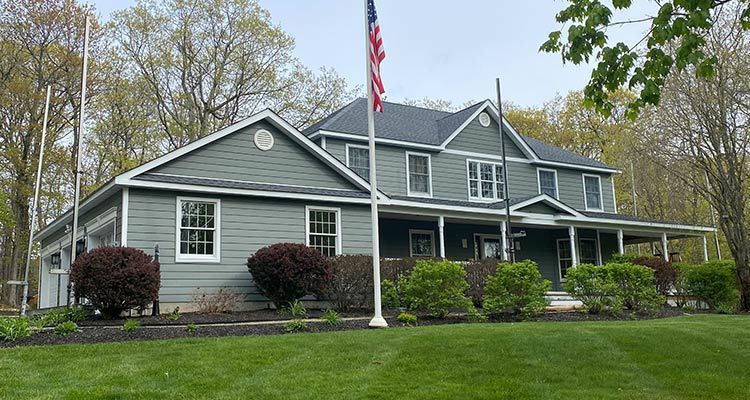 Two-story green building with a porch, American flag, and surrounding trees on a grassy lawn.
