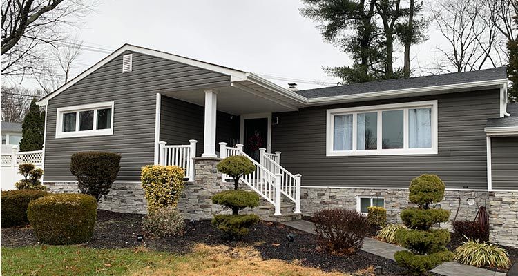 Ranch-style house with gray siding, white trim, stone base, and manicured landscaping under an overcast sky.